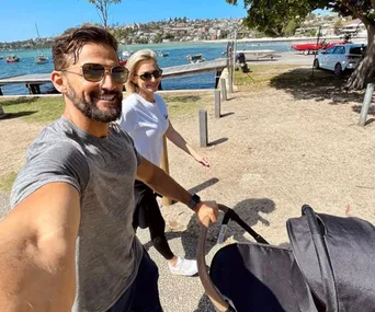 Couple strolling with a stroller by a scenic waterfront on a sunny day, boats and shoreline visible in the background.