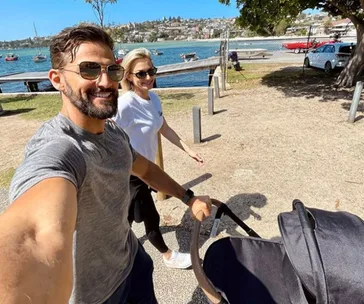 Couple strolling with a stroller by a scenic waterfront on a sunny day, boats and shoreline visible in the background.