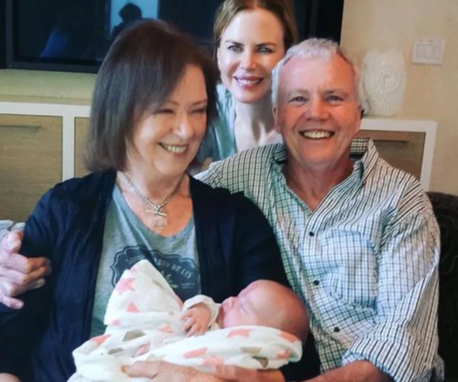 Family photo with a smiling couple and a baby wrapped in a blanket, with another smiling individual in the background.
