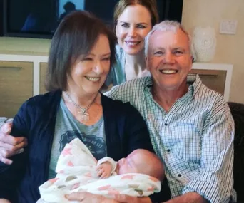 Family photo with a smiling couple and a baby wrapped in a blanket, with another smiling individual in the background.