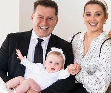 A smiling couple with a baby wearing a white outfit and headband, held by the man in a suit and tie.