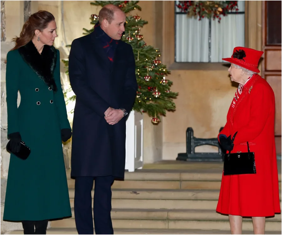 A man and woman in formal coats talk to an elderly lady in red by a festive tree outside a building.