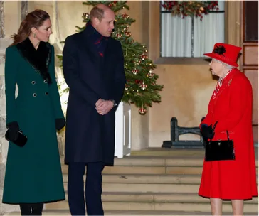 A man and woman in formal coats talk to an elderly lady in red by a festive tree outside a building.