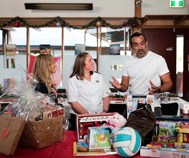 Three people conversing at a table with Christmas decorations and gifts, including a basket, toys, and a volleyball.