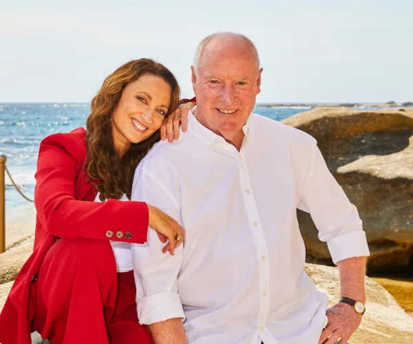 "Two people smiling on a rocky beach, one wearing a red jacket and the other a white shirt, with ocean in background."