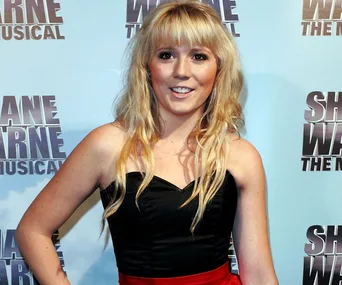 Person with long blonde hair in a black top at "Shane Warne The Musical" event backdrop.