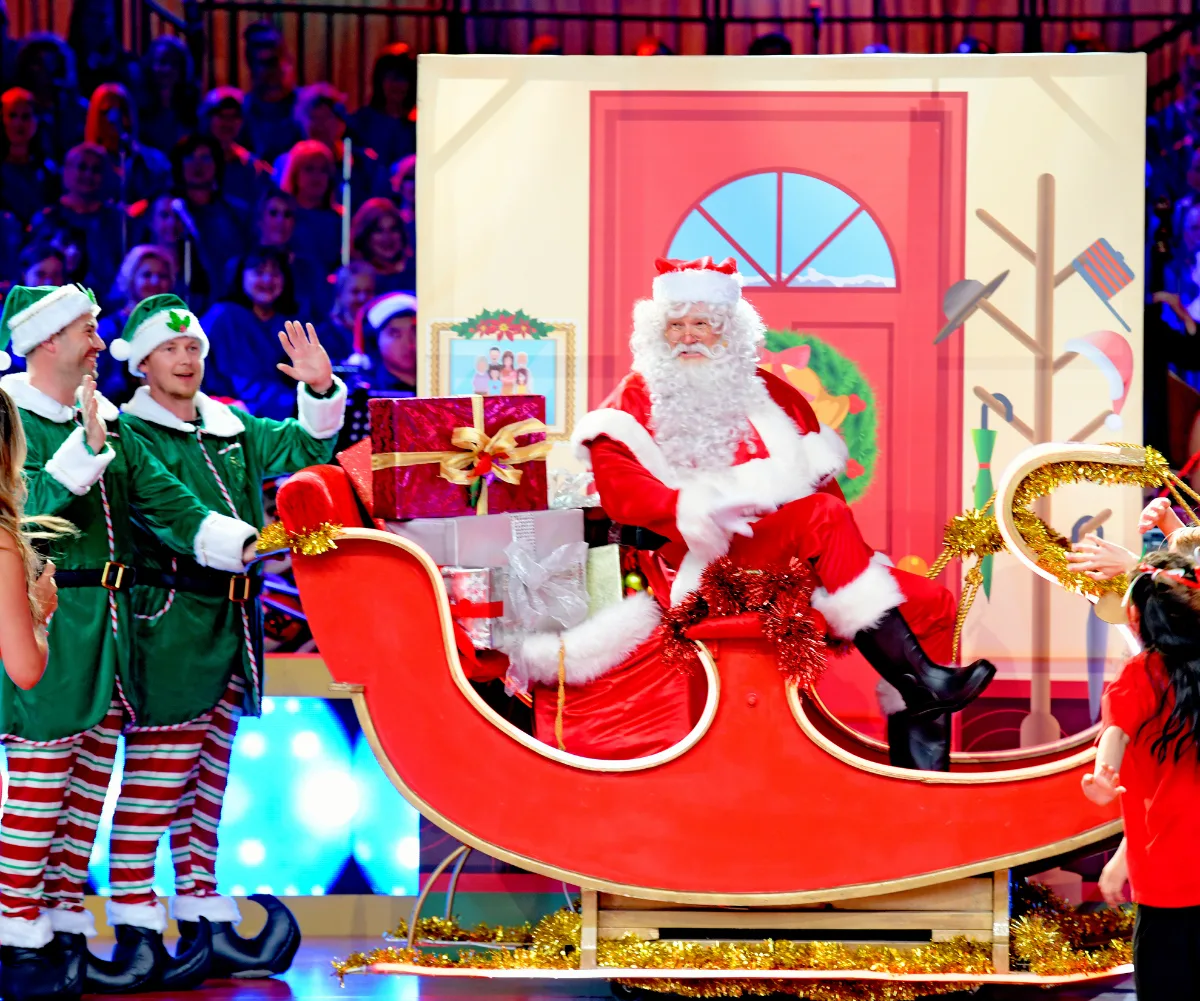 Santa Claus sitting in a sleigh with gifts, surrounded by elves and festive decorations.