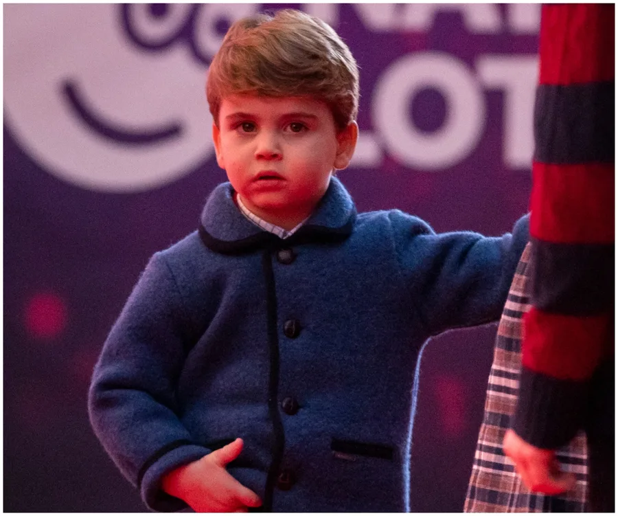 Young child in a blue coat stands against a red background, holding hands with someone off-frame.