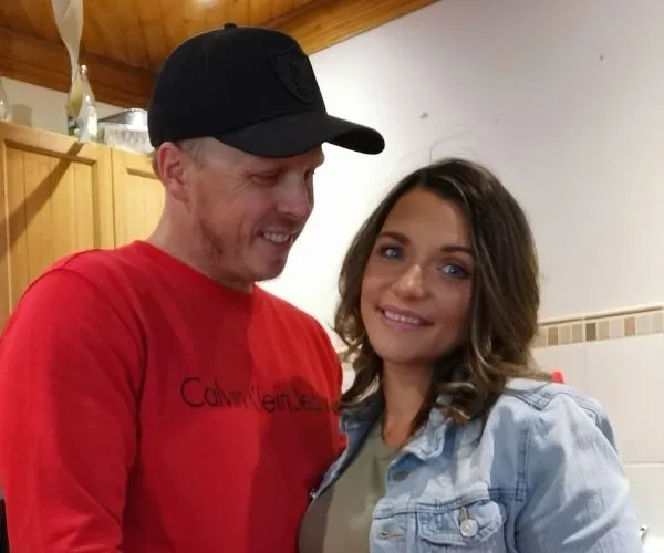 A smiling couple in a kitchen, the woman in a denim jacket, the man wearing a red Calvin Klein sweater and black cap.