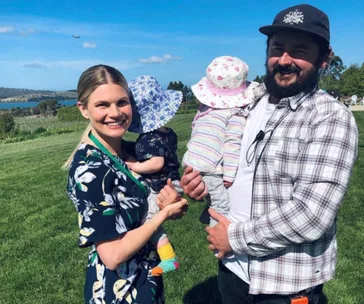 Smiling couple holding two toddlers in sun hats, standing outdoors on a sunny day with a scenic backdrop.