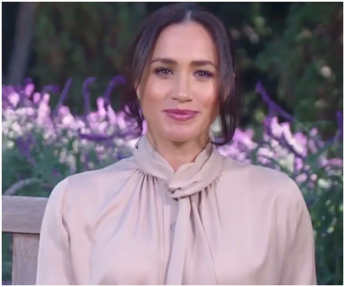 A woman in a light blouse sits outdoors with purple flowers in the background.