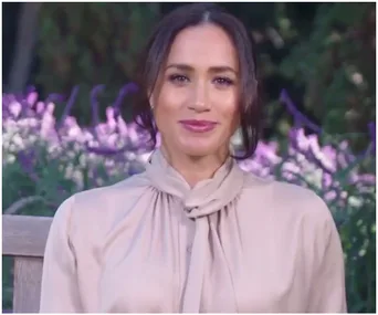 A woman in a light blouse sits outdoors with purple flowers in the background.