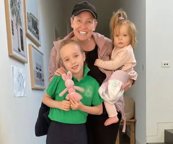 A smiling person with a cap holding a toddler and standing next to a child in a green shirt holding a pink toy.