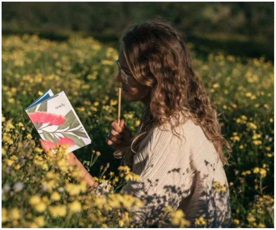 A person sits in a field of yellow flowers, reading a book with a floral cover, holding a pen to their lips.