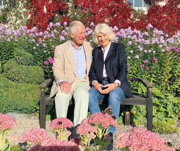 A couple sits on a garden bench surrounded by vibrant flowers, smiling and enjoying the sunny day.