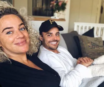 A woman with curly hair and a man wearing a baseball cap sit on a couch, smiling at the camera in a cozy room.