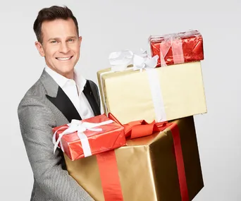A person in a silver suit holding a stack of festive, gift-wrapped presents, smiling at the camera.