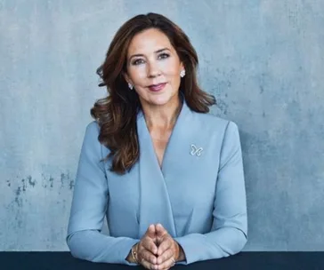 Princess Mary in a blue suit, sitting with hands clasped, against a gray textured background.