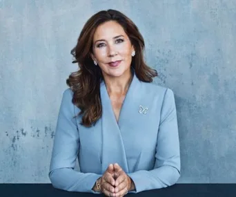 Princess Mary in a blue suit, sitting with hands clasped, against a gray textured background.