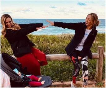 Two women jokingly pose on a wooden fence by the beach, with a stroller and ocean in the background.