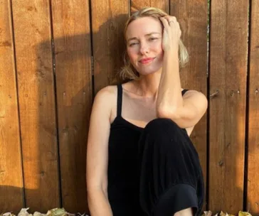 A woman in a black outfit sits against a wooden fence, smiling and resting her hand on her head.