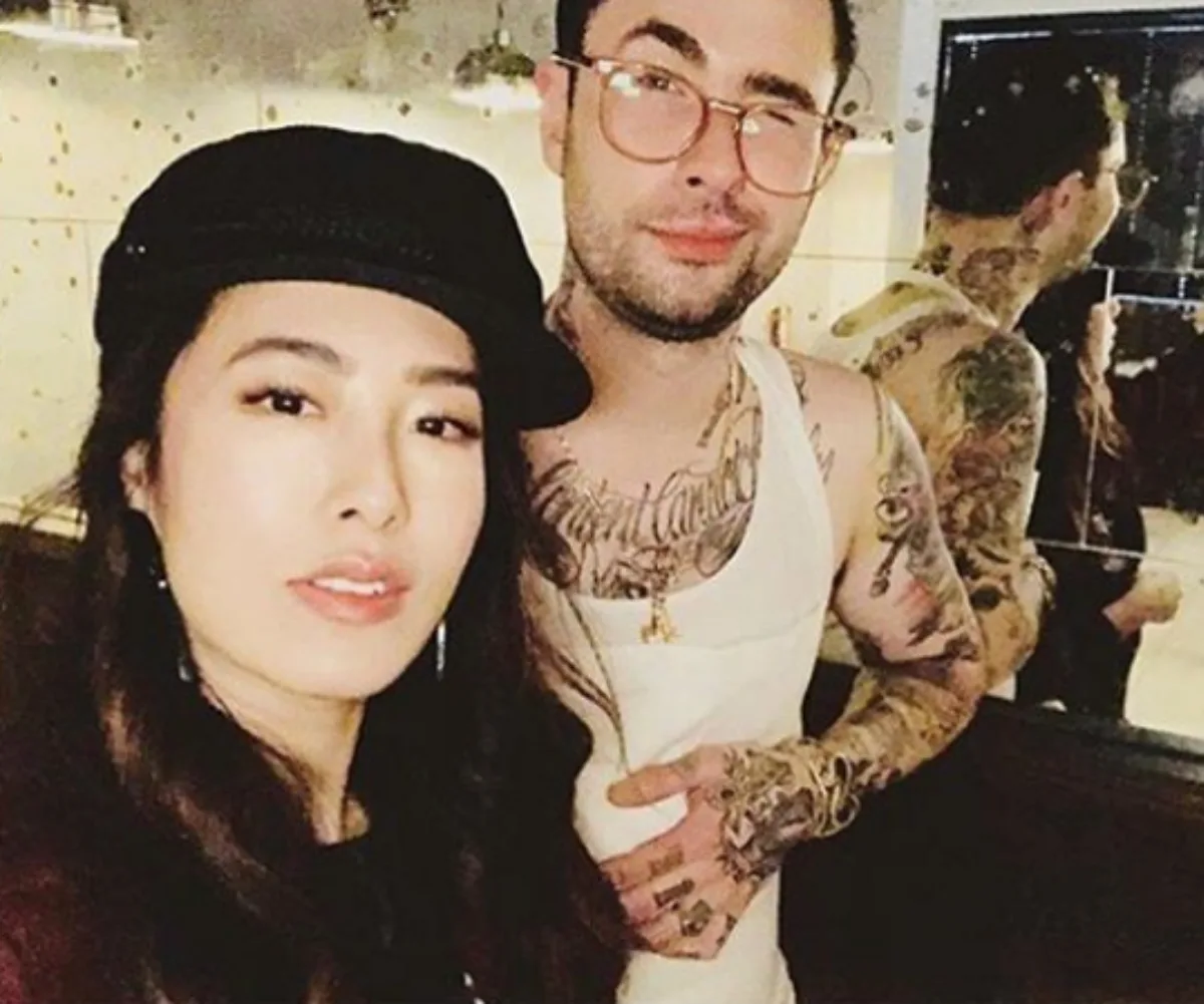 Two individuals posing, one wearing a black hat, the other with tattoos and a white tank top, against a mirrored backdrop.