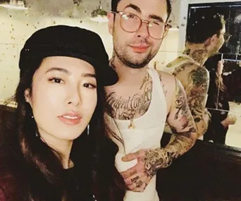 Two individuals posing, one wearing a black hat, the other with tattoos and a white tank top, against a mirrored backdrop.