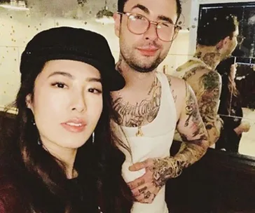 Two individuals posing, one wearing a black hat, the other with tattoos and a white tank top, against a mirrored backdrop.