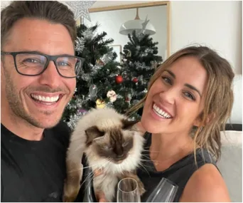 Couple smiling with a fluffy cat in front of a decorated Christmas tree.