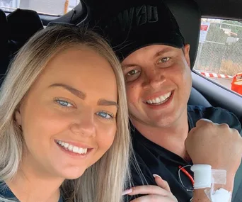 A couple smiling in a car, with one wearing a hat and showing a medical bandage on their wrist.
