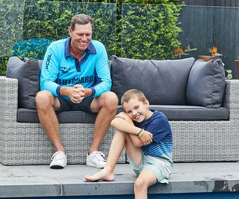 Man in a lifeguard shirt sits on a sofa, smiling with a young boy sitting by the pool's edge, both looking happy.