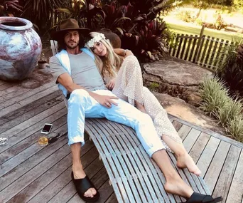 A couple relaxes on wooden deck chairs, surrounded by lush plants, with a drink and phone on the ground nearby.