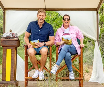 Two people sit on chairs outdoors, smiling, with I'm a Celebrity cards and a buzzer between them.