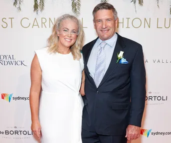 A woman in a white dress with a netted headpiece and a man in a suit with a boutonnière, smiling at an event.