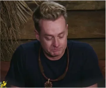A person with closed eyes, wearing a navy shirt and necklace, sitting against a wooden background with foliage.