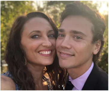 Two people smiling closely, outdoors with greenery in the background.