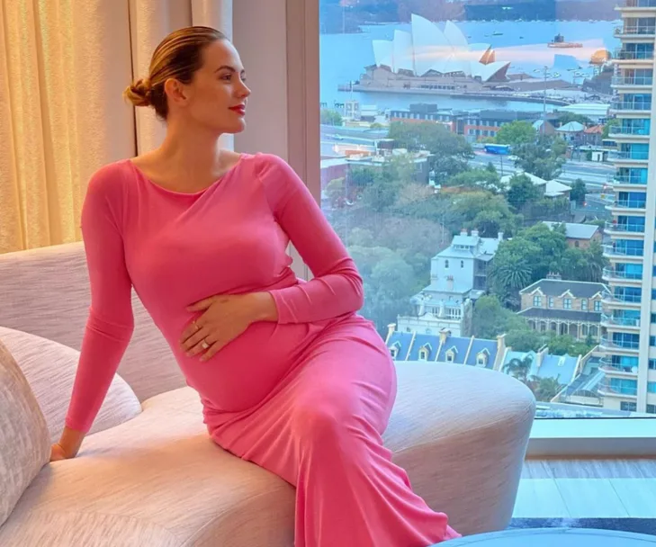 Pregnant woman in pink dress sitting by a window with Sydney Opera House in the background.