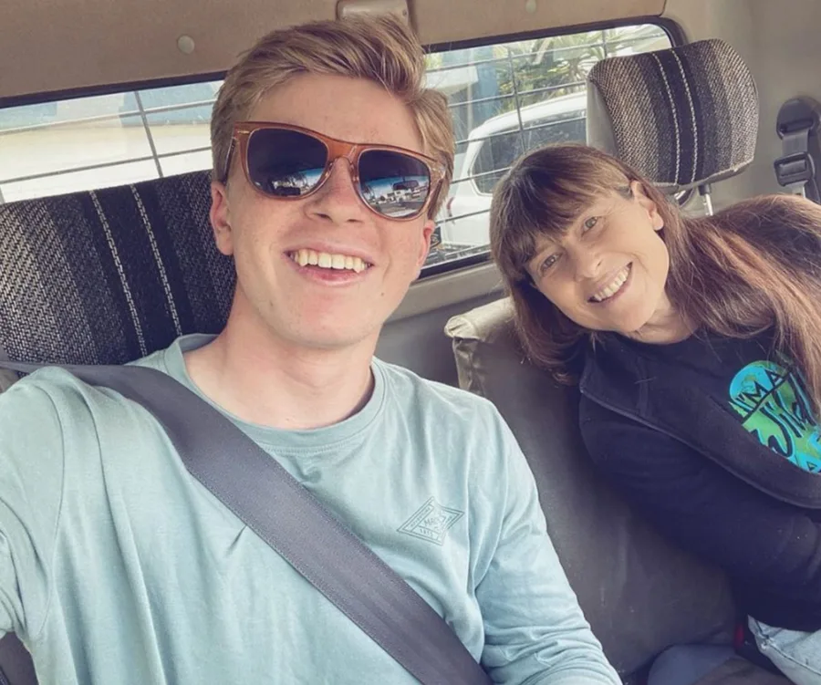 Two people smiling in a car, one wearing sunglasses and the other with long hair, both looking at the camera.