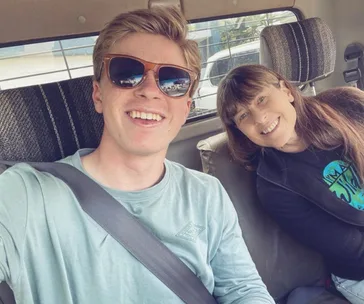 Two people smiling in a car, one wearing sunglasses and the other with long hair, both looking at the camera.