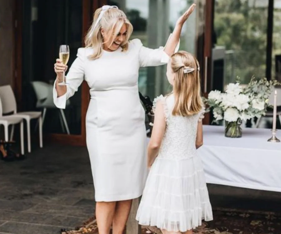 Woman in white dress holding a glass, joyfully talking to a young girl at a wedding, with flowers in the background.