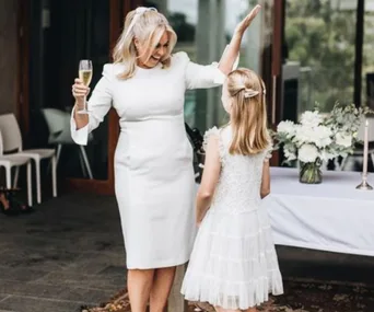 Woman in white dress holding a glass, joyfully talking to a young girl at a wedding, with flowers in the background.