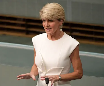A person with short blonde hair, wearing a sleeveless white dress, speaks in a formal setting with a focused expression.