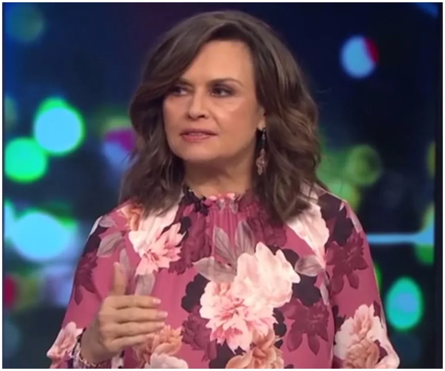 A woman in a floral blouse speaks on a TV show set with colorful blurred lights in the background.