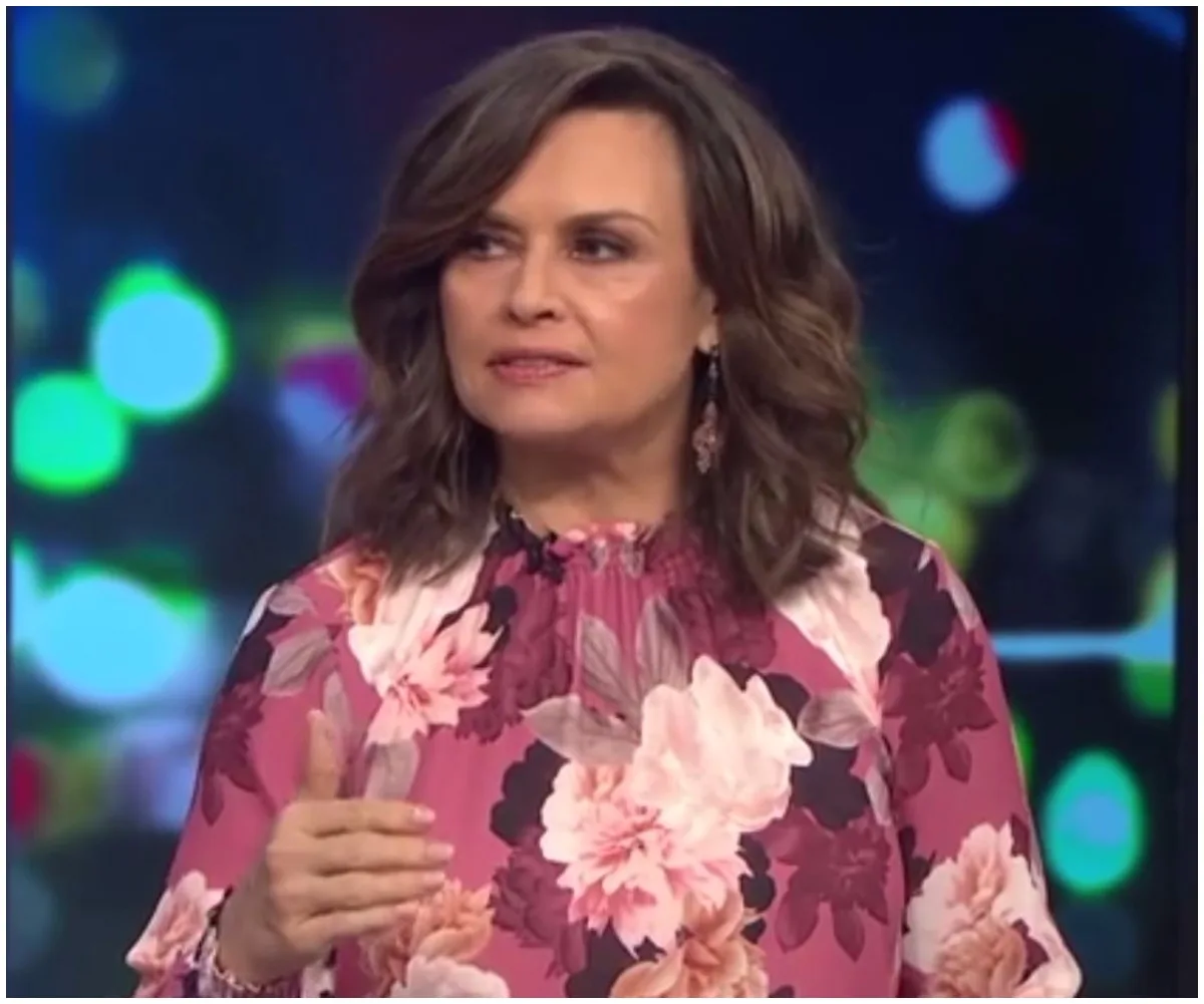 A woman in a floral blouse speaks on a TV show set with colorful blurred lights in the background.