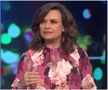 A woman in a floral blouse speaks on a TV show set with colorful blurred lights in the background.