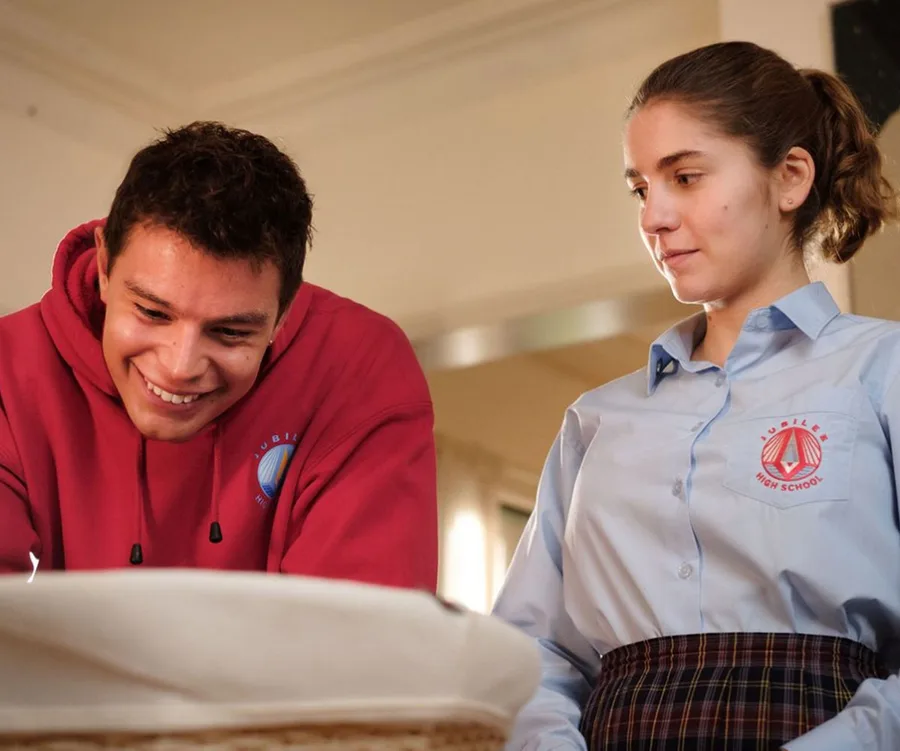 Two young people in school uniforms smile at something off-camera; one wears a red hoodie, the other a blue shirt.
