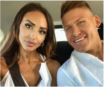 A couple smiling while seated in a car, with the woman wearing a white top and cross necklace.