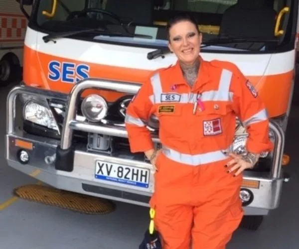 Woman in orange SES uniform smiling in front of an SES vehicle inside a garage.