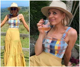 A woman in a wide-brim hat and colorful top smiles outdoors, holding a wine glass, with scenic background.