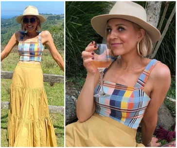 A woman in a wide-brim hat and colorful top smiles outdoors, holding a wine glass, with scenic background.